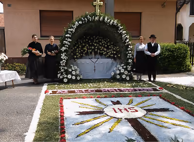 Virágokkal díszített oltár kereszttel, körülötte hagyományos viseletbe öltözött emberekkel, egy fűvel borított vallási jelkép mellett.