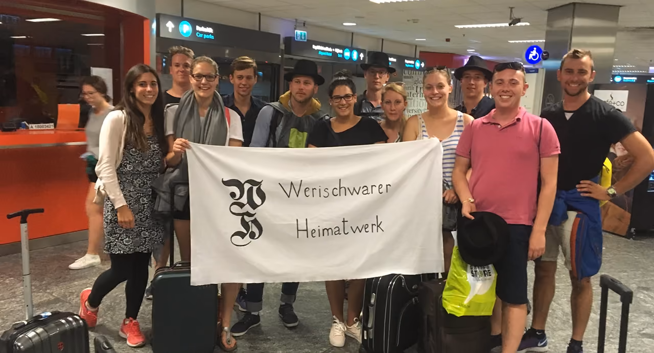 Emberek egy csoportja „Werischwarer Heimatwerk” feliratú fehér transzparenst tartva, poggyászokkal egy repülőtéren.