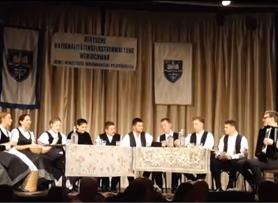 Hagyományos ruházatot viselő emberek csoportja ül asztalok mögött a színpadon, felettük transzparensekkel.