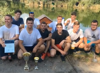 Fiatal férfiak egy csoportja a szabadban, vízparton, trófeákkal és egy oklevéllel, egy elért eredményt ünnepelve.