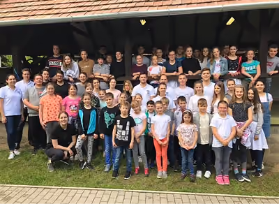 Gyerekek és felnőttek csoportképe egy faépület előtt, a szabadban.