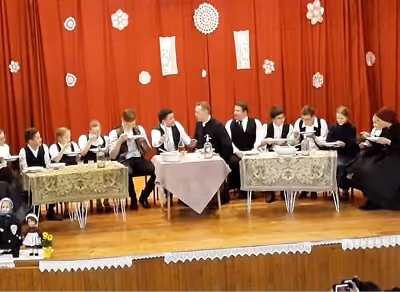 Hagyományos ruhába öltözött gyerekek ülnek három asztalnál a színpadon, vörös függönyös háttér előtt.