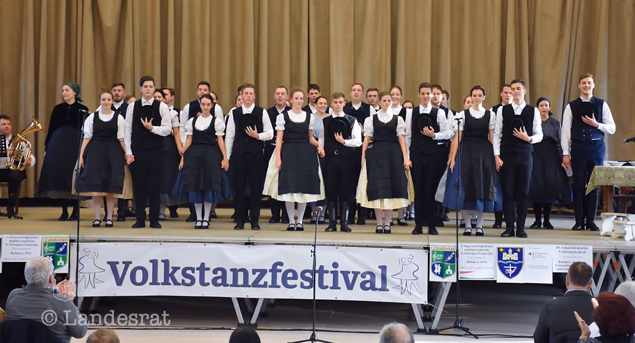A csoport teljes létszámban népviseletben áll a Volkstanzfestival színpadán, előttük a fesztivál feliratú molinóval és mikrofonokkal.