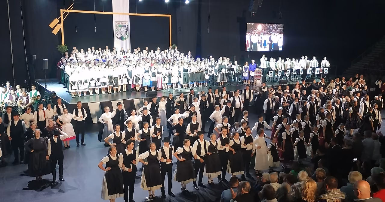 Több száz népviseletes táncos egy nagy rendezvénycsarnokban, a színpadon kórussal, középen körtáncot járó csoportokkal, körülöttük a közönséggel.