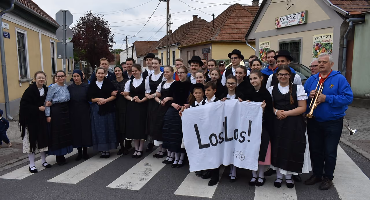 A csoport népviseletben egy zebrán állva, középen „Los! Los!” feliratú transzparenssel, mellettük trombitás zenész, a háttérben falusi házak.