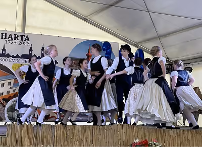 Táncosok sváb népviseletben egy rendezvénysátor színpadán, mögöttük a 300 éves Harta jubileumának molinója.