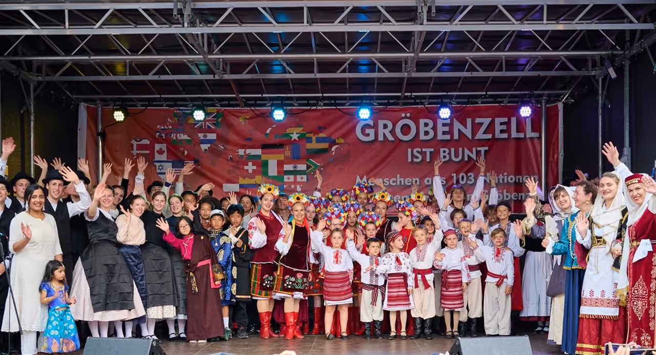 Sok nemzet népviseletes csoportjai integetnek együtt a gröbenzelli fesztivál színpadán, mögöttük a „Gröbenzell ist bunt” feliratú zászlós molinó.